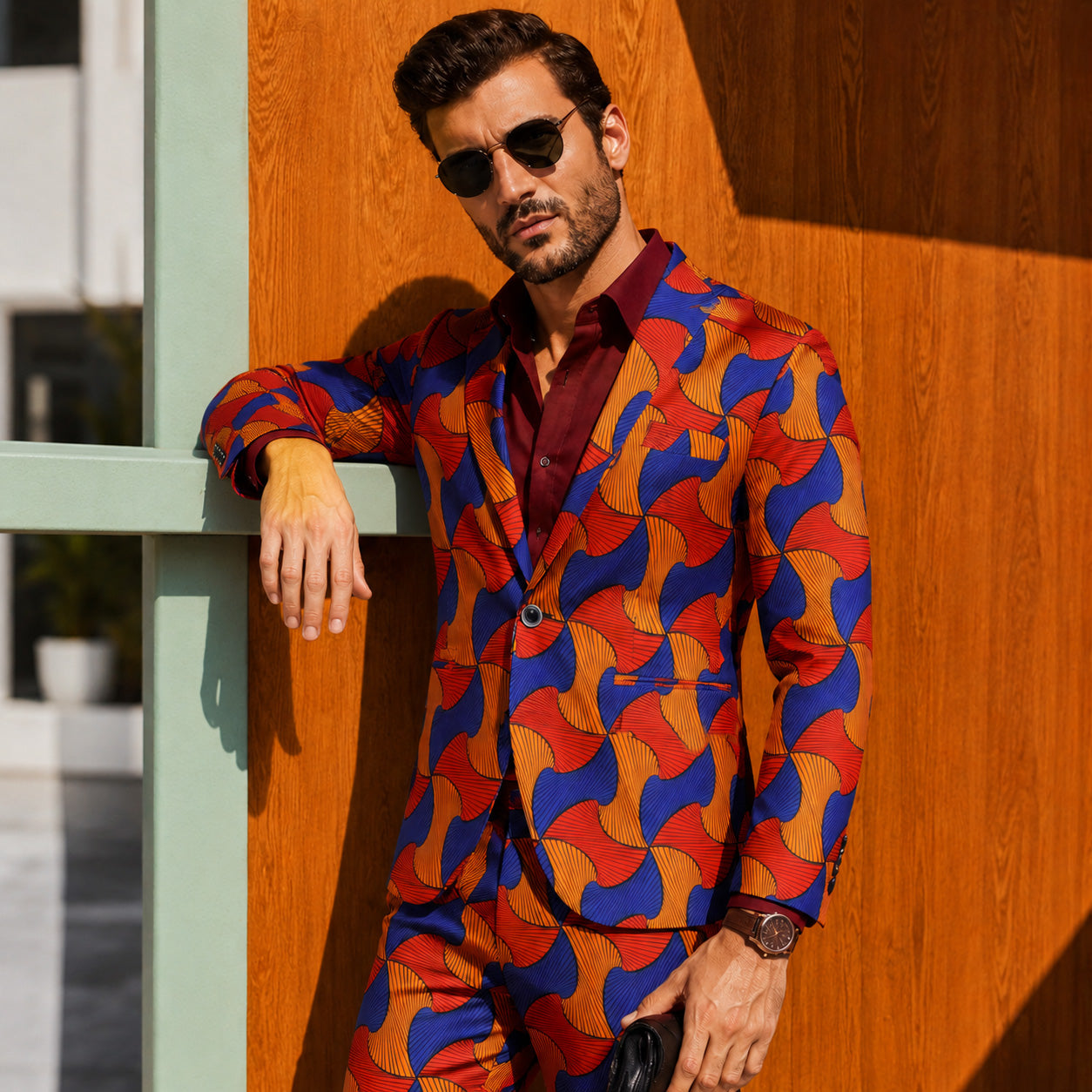 Side view of a stylish man leaning against a wooden wall, wearing an African Ankara print suit blazer with a burgundy button-down shirt and sunglasses.