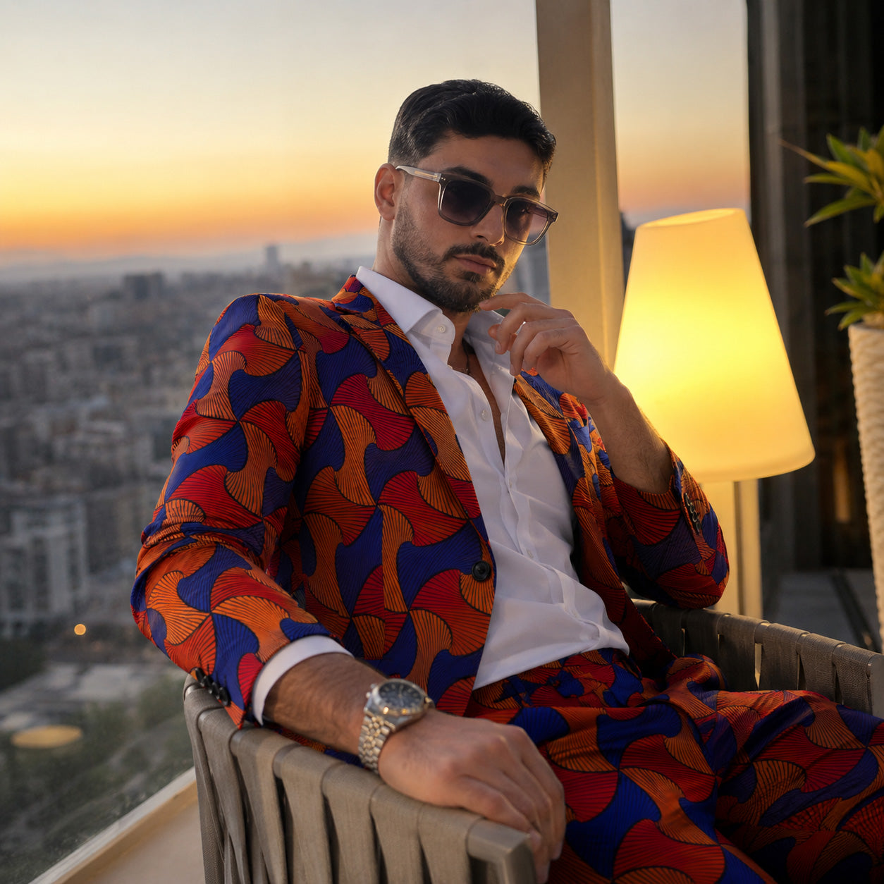 A handsome man sitting on a rooftop terrace at sunset, wearing an orange and blue African pattern men's suit jacket for an upscale evening party.