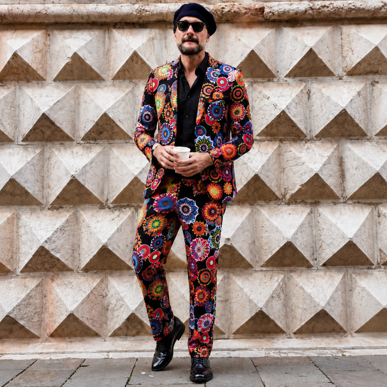 A man wearing a premium slim-fit mens suit with a vibrant colorful artistic mandala print, styled with a black shirt and dark beret against a geometric stone wall background.