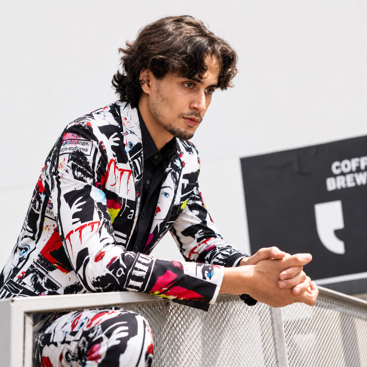 Model in an artistic graffiti patterned suit and white t-shirt sitting on a black spiral staircase, looking forward.
