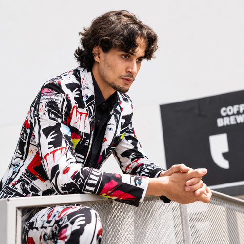 Model in an artistic graffiti patterned suit and white t-shirt sitting on a black spiral staircase, looking forward.