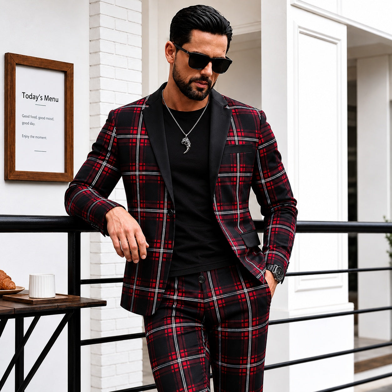 A modern slim-fit men's red and black plaid suit with solid black lapels, worn with a black t-shirt and sunglasses in an upscale outdoor setting.