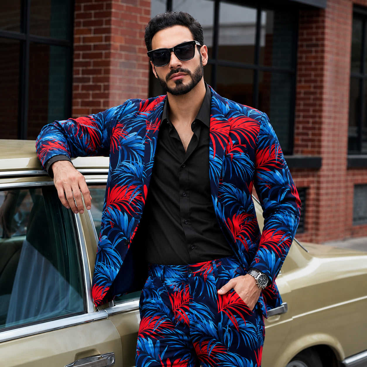 Confident man leaning against a vintage gold car wearing a bold blue and red tropical print blazer and matching trousers with a black shirt and sunglasses.