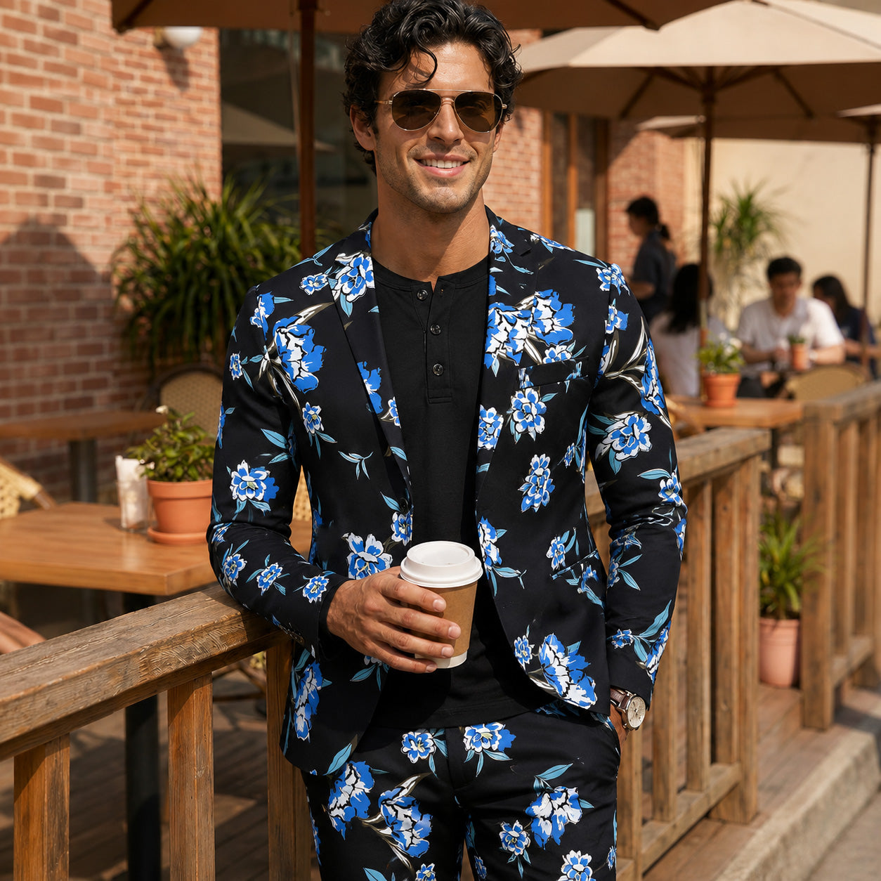 Handsome man wearing Wisholic men's blue and white floral print suit with black inner shirt, holding a coffee cup at an outdoor cafe.