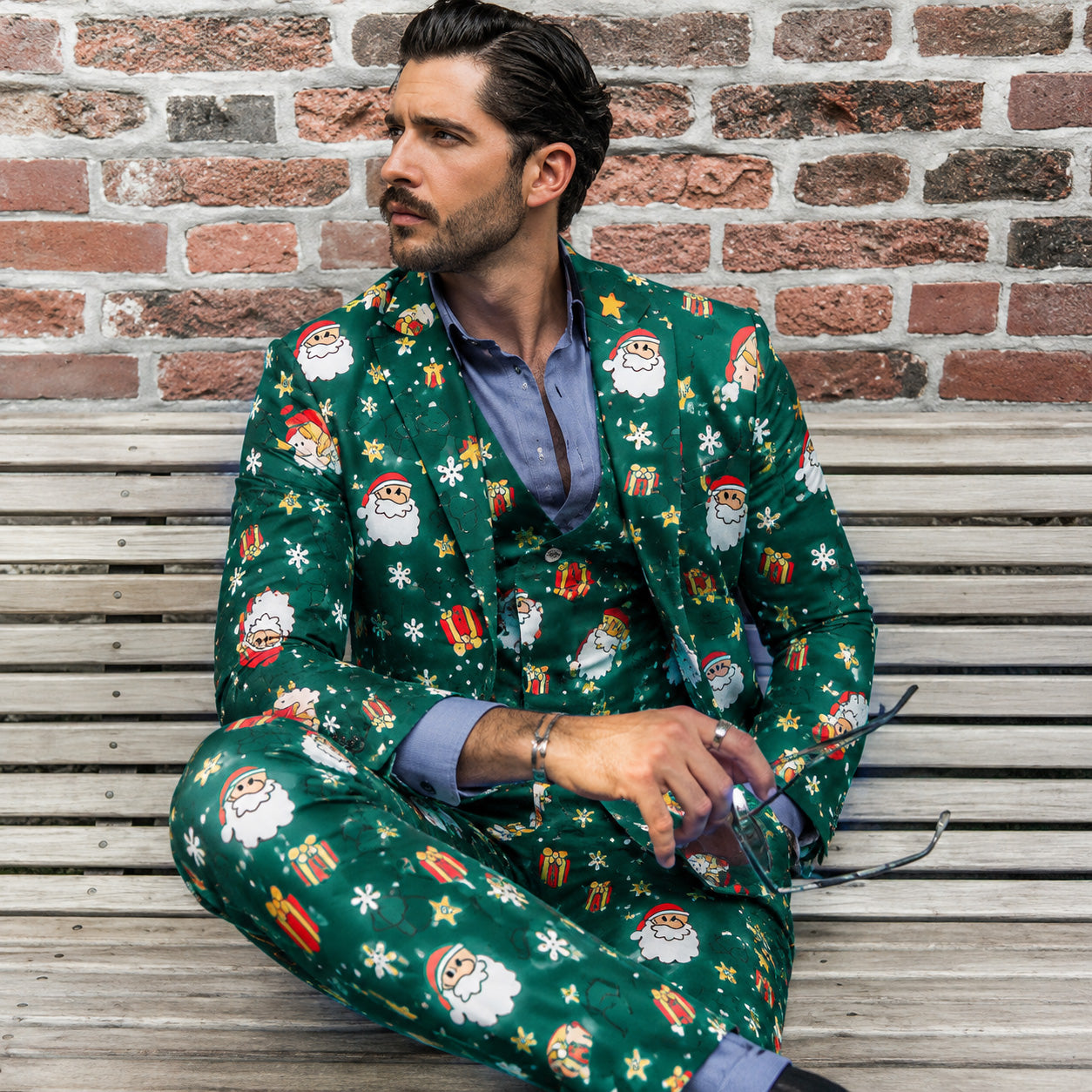 Man sitting on a bench against a brick wall in a WISHOLIC green festive Santa print suit styled with a unbuttoned shirt.