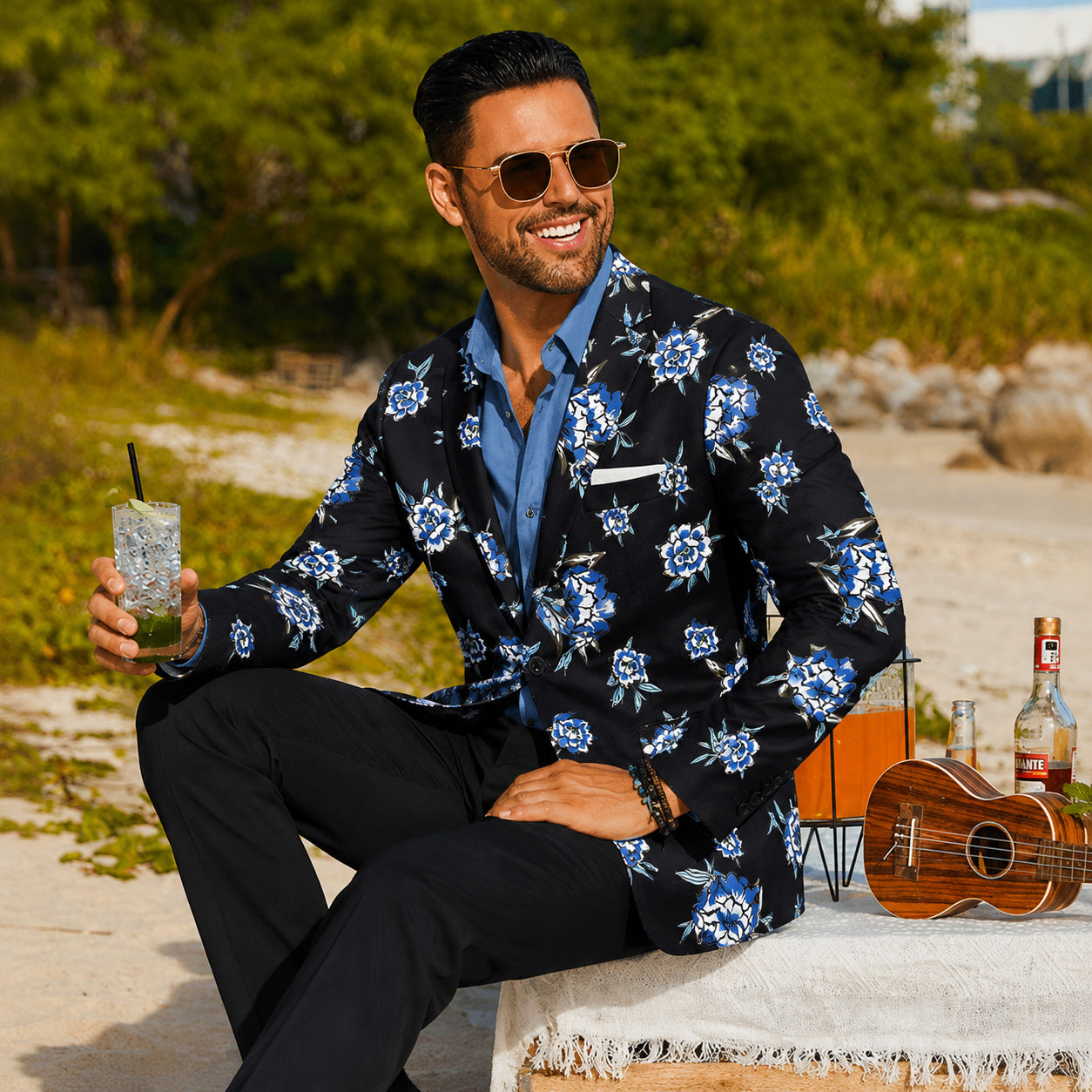 Stylish man in Wisholic blue floral print blazer, light blue shirt, and black dress pants relaxing on a beach vacation.