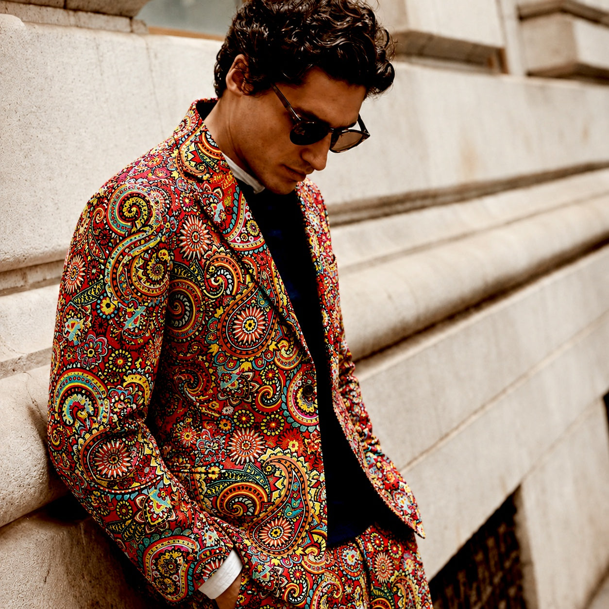 A close-up side profile of a man wearing the Wisholic red paisley artistic printed suit blazer, leaning against a classic stone wall in an editorial street setting.