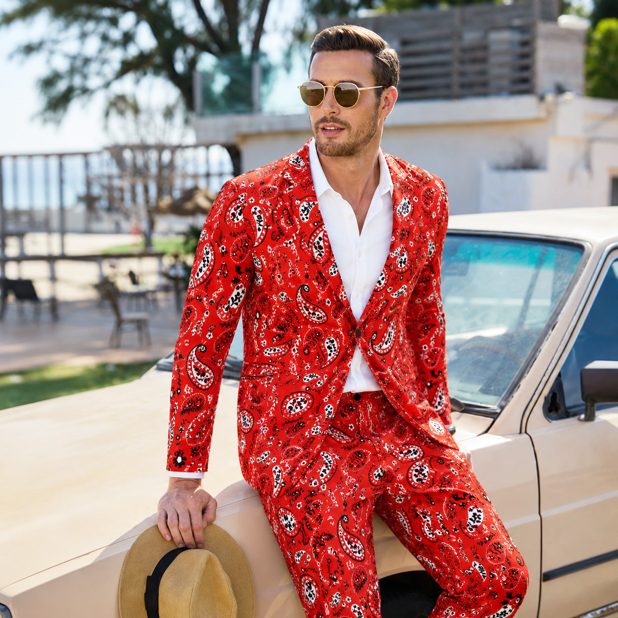 A stylish man in a Wisholic red paisley printed suit leaning on a vintage car, holding a fedora hat.